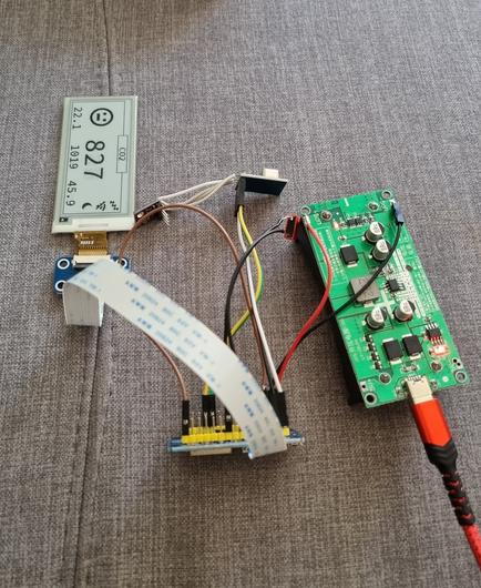 Links oben ist ein eInk-Display mit einem unzufriedenen Emoji, der Zahl 827 und einigen weiteren Zahlen und Symbolen. 
Rechts im Bild eine elektronische Platine. 
In der Mitte ein Mikrocontroller mit diversen Kabeln.