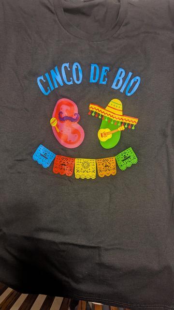 The front of a black T-shirt. It says "Cinco de Bio" and has a mitochondria with a mustache and maraca and a chloroplast with a sombrero and a gitarron