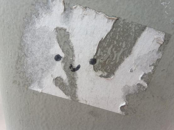 Grauer Ampelmast mit den Resten eines rechteckigen Aufklebers (etwas Papier, sonst Klebefläche). In der Mitte ein kleiner Smiley, wahrscheinlich mit Edding gemalt.