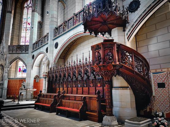 Innenaufnahme der Schlosskirche Wittenberg mit kunstvoll geschnitzter hölzerner Kanzel, hohen gotischen Fenstern mit buntem Glas, historischen Kirchenbänken und verzierten Steinbögen.