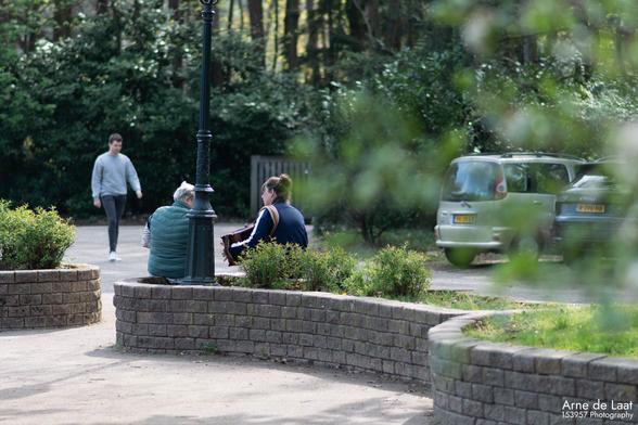 Een foto van twee mensen buiten op een muurtje, gefotografeerd van achteren en een beetje ver weg. Ze spelen harmonica en lijken in interactie met elkaar.