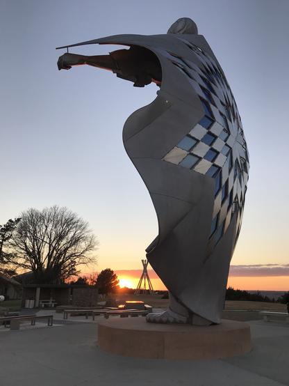 A silver-tone, multi-story statue of a native woman wrapped in a blanket with sunset in a clear sky behind it.