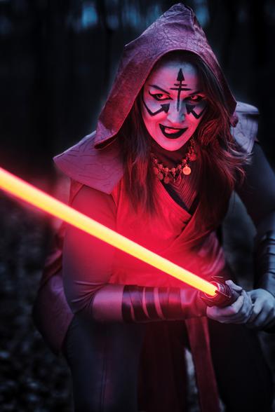 Picture of Lili in a red and black Nightsister inspired Outfit from the Star Wars Universe. She is prowling with her activated red Lightsaber and is licking her lips while looking at her "prey".