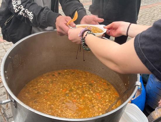 Nad dużym garem z zupą, jest wydawana ona w plastikowej misce do rąk osobie, która trzyma łyżkę.