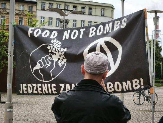 Osoba w czarnej kurtce i czapce stoi przed zawieszonym banerem Food Not Bombs, na którym w górnej części widnieje napis "FOOD NOT BOMBS", poniżej logo Food Not Bombs (pięść skierowana do góry, trzymająca marchewkę) i znak anarchii (litera A w kółku), a na samym dole jest napisane "JEDZENIE ZAMIAST BOMB"