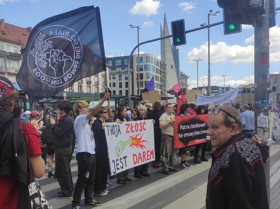 Liczna grupa osób zgromadzona podczas demonstracji 1-majowej. Na zdjęciu szczególnie wyrożnia się osoba trzymająca czarną flagę z logiem Food Not Bombs, otoczonym kołem z napisem "FOOD NOT BOMBS BIELSKO-BIAŁA", a obok parę innych trzymających baner "TWOJA ZŁOŚĆ JEST DAREM", na którym widnieje koktajl mołotowa z etykietą symbolu antify (trzy strzałki skierowane na ukos)
