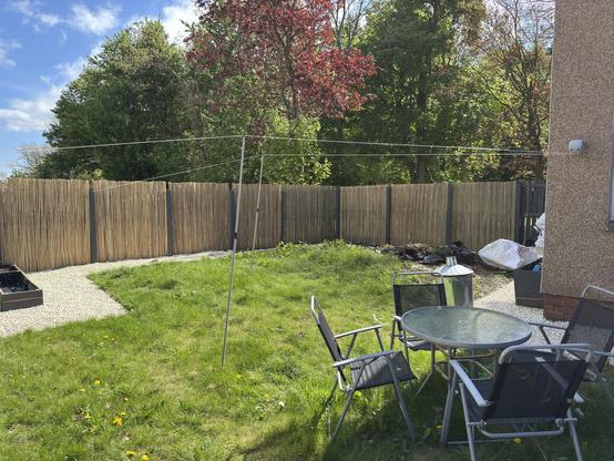 A backyard scene featuring a grassy area with a picnic table and chairs, a clothesline, and a wooden fence. Lush trees are visible in the background, alongside some garden waste in bags. The sky is partly cloudy.