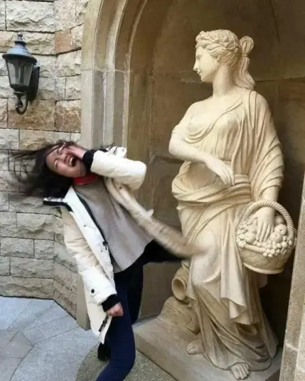 A young woman humorously "slaps" a classical-style statue in an unknown location. The statue, dressed in flowing robes and holding a basket, appears to recoil, creating a playful interaction that blurs the line between art and reality.