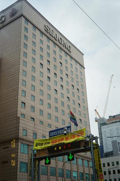 서울 세종호텔 앞 도로표지판에서 고공농성 중인 고진수 동지. 전광판 위에 “윤석열 파면! 세종호텔 해고자 복직!”이라는 현수막이 걸려있고 “세종호텔지부”라고 써있는 무지개 깃발이 나부끼고 있다.