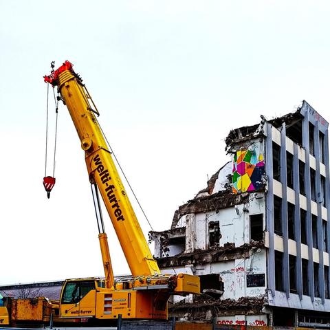 This image depicts a construction scene featuring a yellow crane positioned next to a partially demolished building. The crane is equipped with a hook, ready to assist in the demolition process. The exposed structure shows signs of wear, and a colorful abstract graffito visible on one section of the wall.