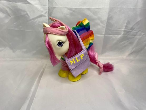 A colorful toy pony with pink hair, wearing a headband and a striped dress, holding a small purple bag labeled "MLP." The background is plain white.