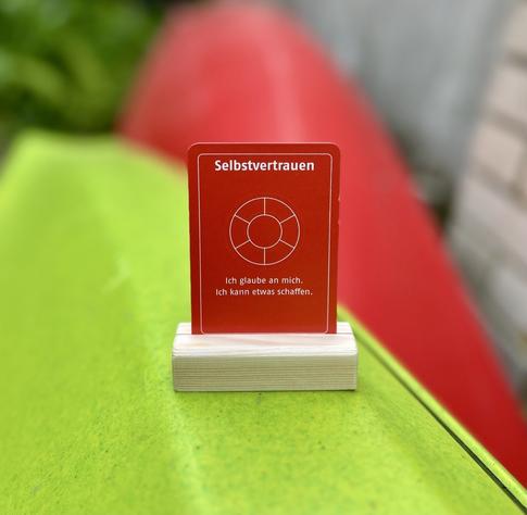 Auf einem grünen Untergrund steht eine rote Wertekreis-Karte mit dem Wert „Selbstvertrauen“ im Holzständer. Darauf: „Ich glaube an mich. Ich kann etwas schaffen.“