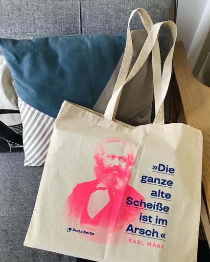 Unsere Marx-Baumwolltasche trägt den Aufdruck »Die ganze alte Scheiße ist im Arsch« in Blau, daneben ist ein aufgerastertes Porträt von Karl Marx in Neonrot zu sehen. Das Zitat stammt von Karl Marx, geschrieben an Friedrich Engels zur Wirtschaftskrise 1857; Quelle: Marx-Engels-Werke Band 29, Seite 239.
