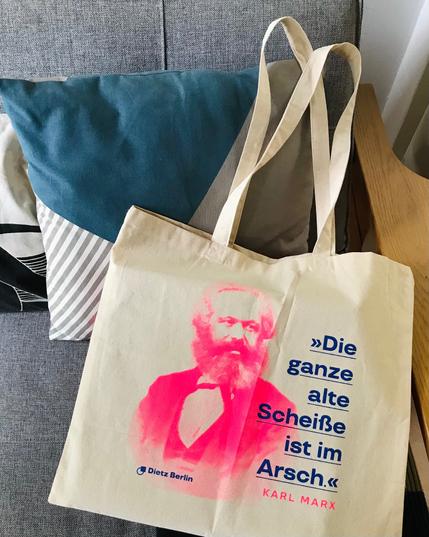 Unsere Marx-Baumwolltasche trägt den Aufdruck »Die ganze alte Scheiße ist im Arsch« in Blau, daneben ist ein aufgerastertes Porträt von Karl Marx in Neonrot zu sehen. Das Zitat stammt von Karl Marx, geschrieben an Friedrich Engels zur Wirtschaftskrise 1857; Quelle: Marx-Engels-Werke Band 29, Seite 239.