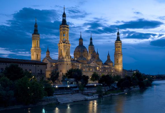 The Nuestra Señora del Pilar Basilica in Zaragoza Spain, shot during the blue hour at dusk, while the basilica is illuminated by yellowish spotlights under a deep blue partly cloudy sky.