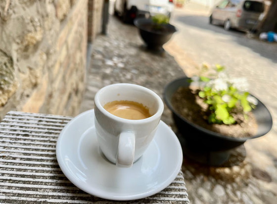 A small cup of eswpresso placed on a pedestal in focus. Walls, cobblestone street and pots with plants are in bokeh.