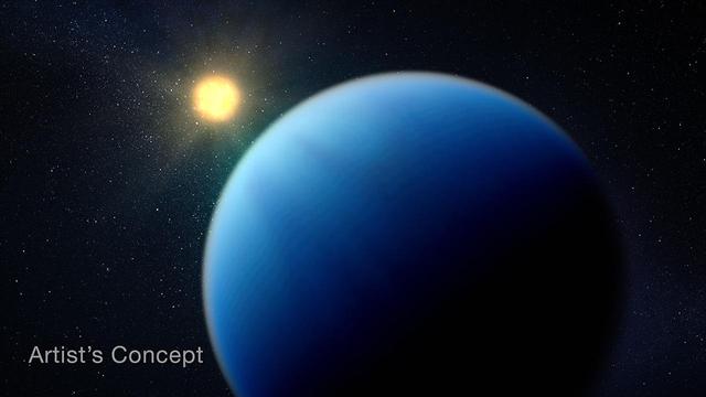 Illustration showing a large fuzzy blue planet in the foreground, and an orange-yellow star, which appears much smaller, in the background. The side of the planet facing the star is lit, and the side facing away is dark. The boundary between the lit and dark sides is fuzzy. The atmosphere is almost homogeneous in color and texture, with extremely subtle variations and no sign of a surface. The black background of space is scattered with tiny white points of light. The words “Artist’s Concept” is in the lower left corner.