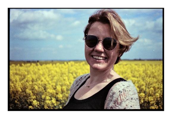 Smiling woman wearing sunglasses, standing in a blooming yellow rapeseed field under a partly cloudy sky, captured on expired Fujicolor 200 film and self-developed, giving the image a grainy, vintage look with magenta shifts and soft contrast.