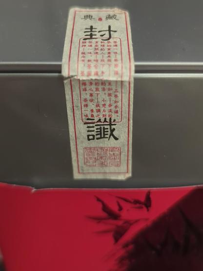 corner of tea tin with broken paper seal displaying nicely set chinese characters. i suspect it shows the variety, shelf name, a description, and processor/company/teamaster.