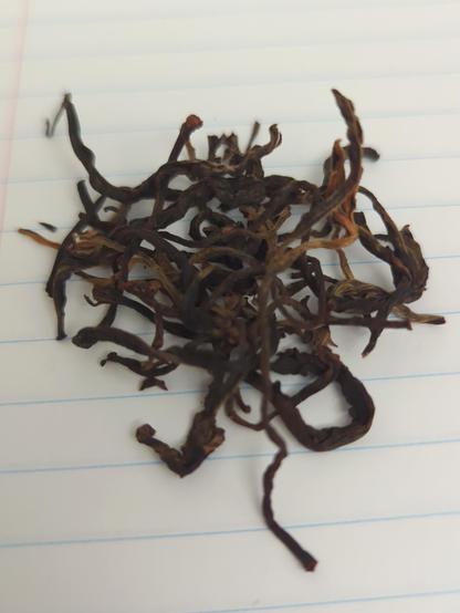 poor photo of dry processed red tea leaves on lined paper, showing some of the roasting level and quality of picked tea buds. appearance is mostly dark greenish-brown mixed with lighter colors of yellow-red (rustlike) for newer buds. 
yummy yummy yummy