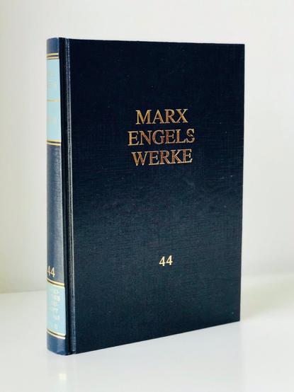 Dreidimensional auf dem Bürotisch stehend fotografiert: Band 44 der Marx-Engels-Werke, Hardcover, in charakteristischem Dunkelblau mit Goldprägung und hellblauen plus goldenen Streifen auf dem Buchrücken.