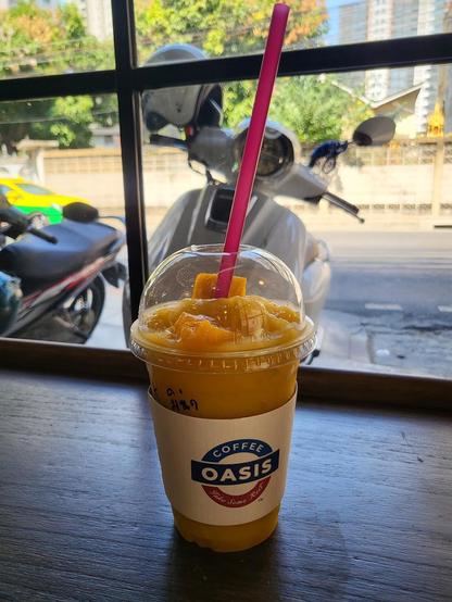 A mango smoothie with a pink straw sits on a table in front of a window. The smoothie is in a plastic cup with a "Coffee Oasis" logo. Outside the window are parked scooters and buildings.