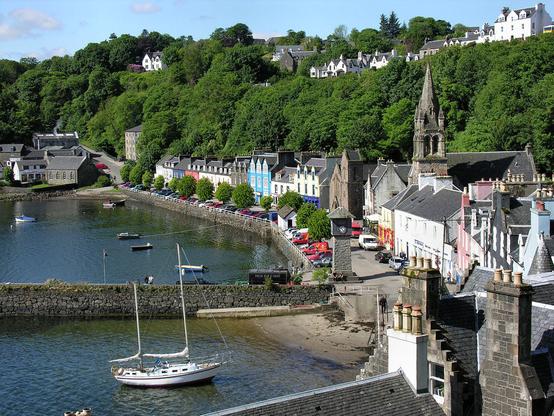 Tobermory on the Isle of Mull. The image shows a view from above one end of a village wrapped around a bay. There are rooftops in the foreground and then the seafront row of buildings continues around to the right and then left, facing left out over a harbour with a stone pier. There are trees behind these buildings, with another row at high level on the upper right, on the ridge line. The scene is in sunshine.