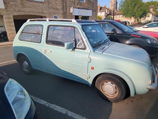 A side view of a light blue Nissan Pao, a small 3-door hatchback with lines moulded into the doors and bonnet reminiscent of a Citroen H-van. It's in a car park with a church in the distance. The paint is practically matte finished, presumably after years of not being polished, yet it looks oddly cared for, considering it's 35-odd years old. The side rear windows are horizontally hinged in the middle, to add to the weird old car vibe.