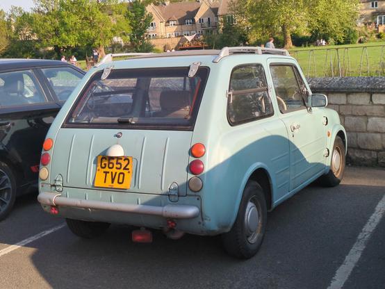 A rear three-quarter view of a light blue Nissan Pao, a small 3-door hatchback with lines moulded into the doors, boot and bonnet, reminiscent of a Citroen H-van. It's in a car park. The paint is practically matte finished, presumably after years of not being polished, yet it looks oddly cared for, considering it's 35-odd years old. The side rear windows are horizontally hinged in the middle, to add to the weird old car vibe.