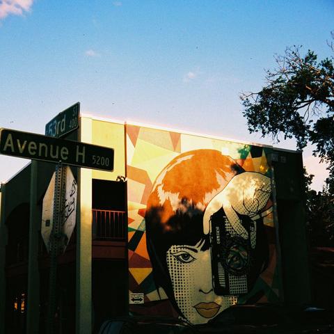 Photograph of the wall of a building with a mural of a person taking a photograph.  
In the foreground is a street sign for 53rd and Avenue H. Strong halation is visible on the top edge of the building.