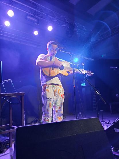 Teacozy op het podium. Hij is in zijn eentje. Hij heeft een wit t-shirt en witte broek met kleurige tekeningen van beestjes. Hij speelt een acoustische gitaar met stickers er op die hij hoog heeft hangen. Naast hem op een kruk liggen een mondharmonica en een versterkertje voor zijn telefoon, die hij ook soms voor een backing track gebruikt.
