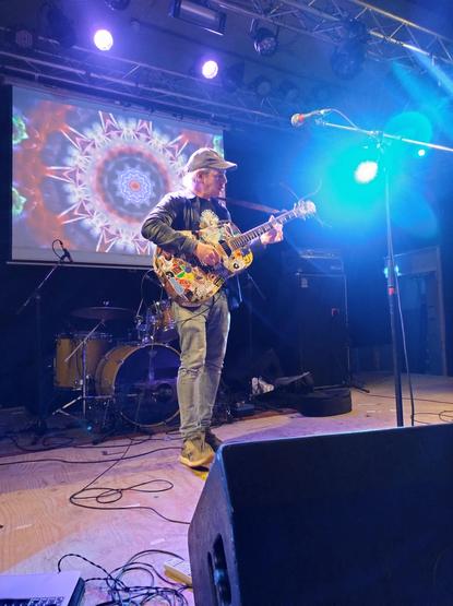 Jeffrey Lewis in zijn eentje op het podium, met volledig met stickers beplakte acoustische gitaar. Jeffrey heeft een leren jasje, t-shirt met een comic figuur er op,  een spijkerbroek en een petje