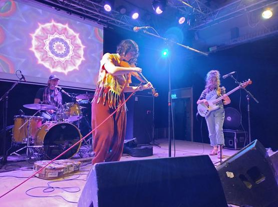 De violiste en bassiste van The Voltage. De violiste heeft een wijde rode broek en een psychedelisch bedrukt oranje t-shirt met rafels. De bassiste heeft een tuinbroek, gestreept t-shirt en opgestoken haar