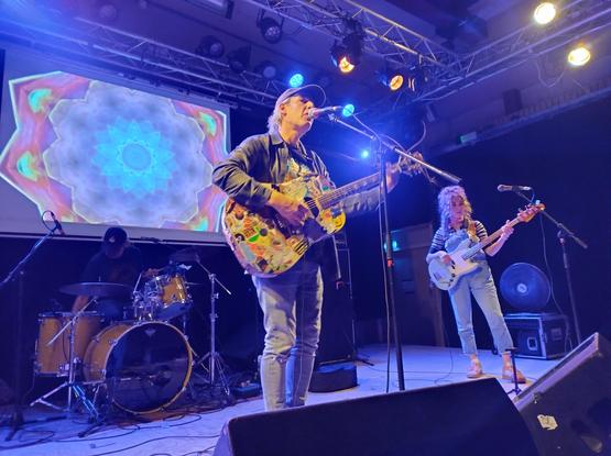 Jeffrey Lewis en de bassiste op het podium. Op de achtergrond de drummer en psychedelische patronen op een scherm.