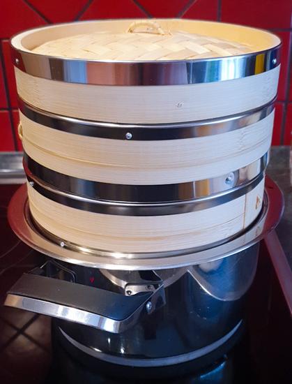 Das Bild zeigt einen Dampfgarer aus Bambus, der auf einem Metalltoptopf steht. Es handelt sich um ein traditionelles, rundes Dämpfgerät, das aus mehreren stapelbaren Bambuskörben besteht. Die Körbe sind mit silberfarbenen Metallringen verstärkt, die zusätzliche Stabilität geben. Der Deckel ist ebenfalls aus Bambus und hat ein kleines Griffstück aus Bast in der Mitte.

Der Dampfgarer steht auf einem glänzenden, silbernen Kochtopf, der auf einem schwarzen Induktions- oder Cerankochfeld platziert ist. Im Hintergrund sind rote Wandfliesen mit schwarzen Fugen zu sehen, was dem Bild einen warmen, traditionellen Küchencharakter verleiht.