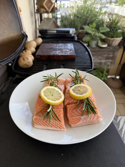 Das Bild zeigt zwei Stücke frischen Lachs auf einem weißen Teller, die mit einer Zitronenscheibe und Rosmarin garniert sind. Der Lachs ist rosa und saftig, und die Zitronenscheiben liegen symmetrisch auf den beiden Stücke. Im Hintergrund ist ein Grill zu sehen, auf dem eine weitere Zutat, möglicherweise Kartoffeln, grillt. Der Grill befindet sich auf einem Balkon, der mit Pflanzen und einem Blick auf die Umgebung geschmückt ist. Die gesamte Szene vermittelt ein Gefühl von frischer Küche und gemütlicher Grillparty.