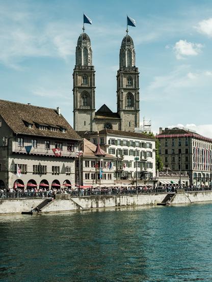 Scenic daytime view of the Grossmünster church with its twin towers rising above historic buildings in Zurich, Switzerland, as crowds gather along the Limmat River under a blue sky with scattered clouds.