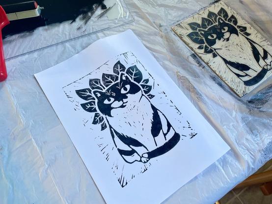 A print of a stout tuxedo cat bsurrounded by basil leaves, next to the cut print block