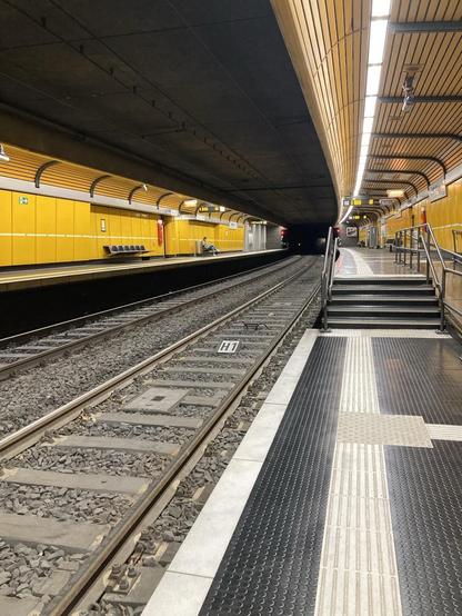 Foto einer U-Bahn Haltestelle vom Trambahnsteig aus fotografiert in Richtung U-Bahnsteig. 5 Stufen führen dort hinauf. Die Wandbekleidung ist sonnengelb gestaltet.