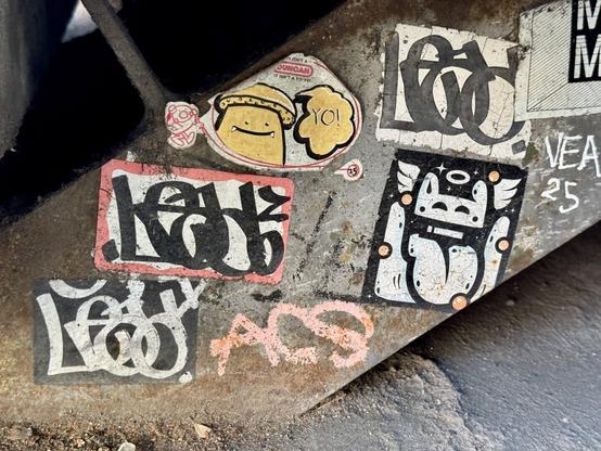 Few graffiti stickers on a metal beam beneath a set of concrete stairs. The stickers have hand drawn art on them by graffiti artists. One in the center has an old Duncan Yo-Yo sticker, but an artist has drawn a mushroom character in yellow and black paint. The mushroom is drawn in Matsu's unique style and says "Yo!". Another awesome hand drawn sticker on the right shows the head of some creature (possibly a bunny) with a big tongue hanging out and angel wings growing out of it's head.