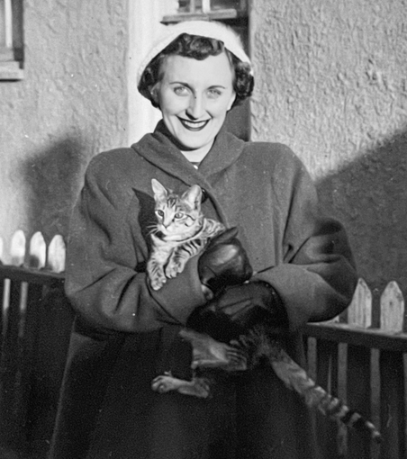 Black and white photo of a light skinned woman in a winter coat, standing outside and smiling at the camera while holding a very cute shorthaired tabby cat in her arms.