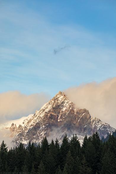 A triangular mountain peak is subtly lit up by the sun as clouds clear around it.  Blue sky with wispy cloud is above the peak, with a foreground of a dark conifer forest.