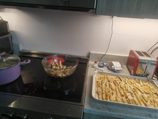 The stages of making homemade French fries happening all at once: a pot of boiling water, a bowl of raw fried, a baking sheet of boiled and drained fries waiting to go in the freezer