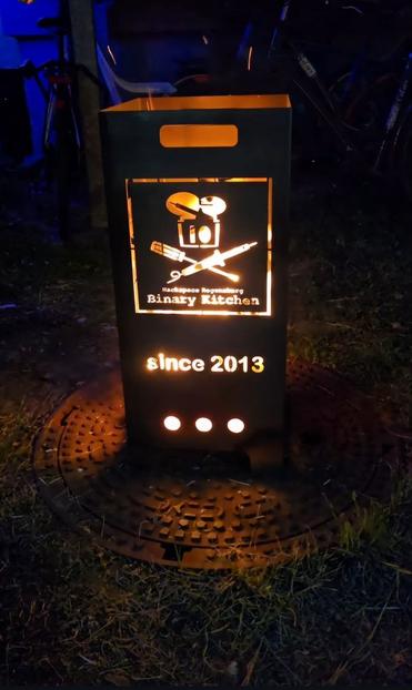 Feuerschale im Dunkeln mit Binary Kitchen Logo sowie "since 2013", Holz darin brennend