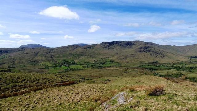 Photo of Connemara landscape, taken from a grassy outcrop, showing a long range of connected hills in the distance across a sheltered valley. A handful of houses and farm buildings can be seen in its crook, amid fields divided by hedges and stone walls. Trees are few except clumps here and there, and some bordering the fields. Behind the hills to the left is Corcóg, a flat mountain pale purple in the distance. The sky is blue with a few white puffy clouds and a lot of thin haze.