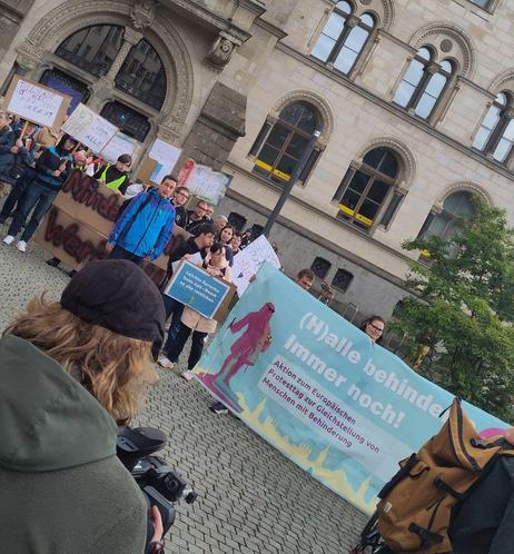 Zahlreiche Menschen protestieren vor einem alten sandsteinfarbenen Prachtbau mit großen runden Fentern und halten Transparente mit Themen wie der Forderung nach Mindestlohn für Werkstatttätige, Teilhabe und Inklusion oder der Verfügbarkeit Leichter Sprache auf Ämtern.