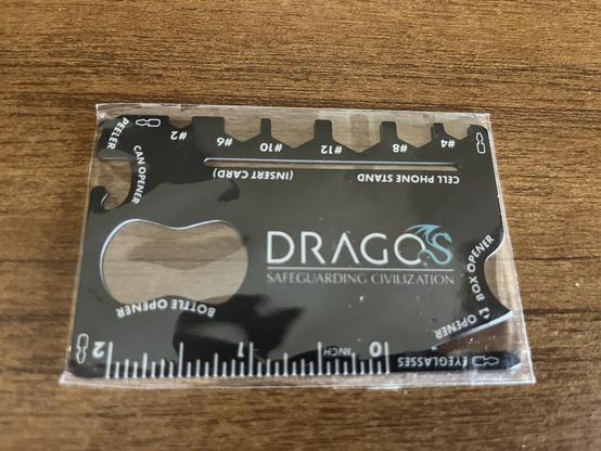 A black, credit-card-sized metal multi-tool branded 'Dragos Safeguarding Civilization', shown inside its clear plastic sleeve on a wooden surface. Visible tools include a bottle opener, can opener, peeler, box opener, ruler, hex wrenches, eyeglass screwdriver, and a cell phone stand slot.