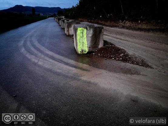 Betonblöcke trennen eine Strasse in den asphaltierten und schlammigen Bereich. Der vorderste Block ist mit gelber Farbe markiert.