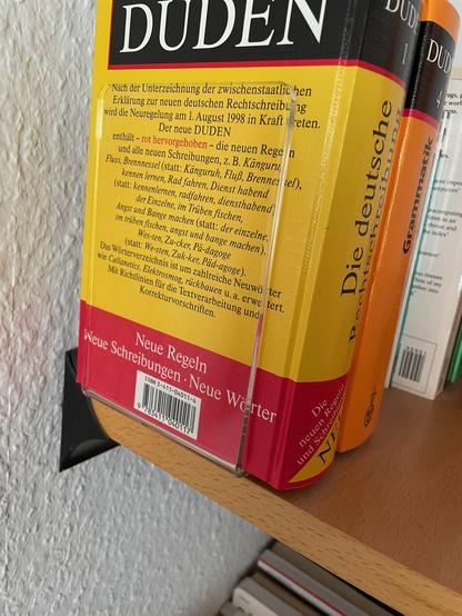 Ein Bücherregal mit Büchern drauf. Es besteht aus einer Regalbrett in Buche-Ausführung und einer an die Wand geschraubten schwarzen Metallschiene, in die das Brett einfach eingeschoben wird.