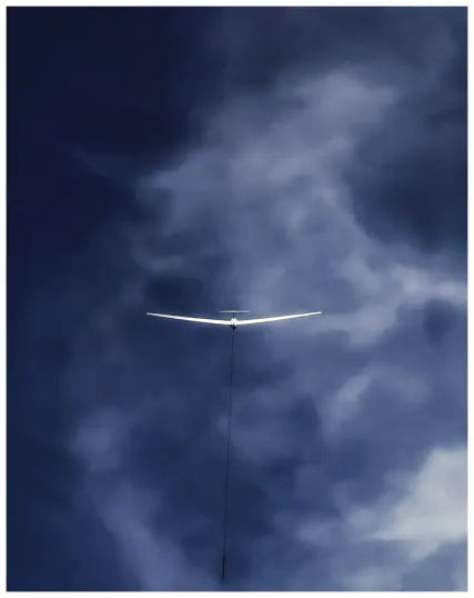 Das Foto zeigt blauen, von weißen Wolkenschleiern durchzogenen Himmel als formatfüllenden Hintergrund. Davor im Bildzentrum die Rückansicht eines vom Sonnenlicht strahlend erhellten weißen Segelflugzeugs, welches an einem Windenseil in den Himmel/die Höhe gezogen wird.